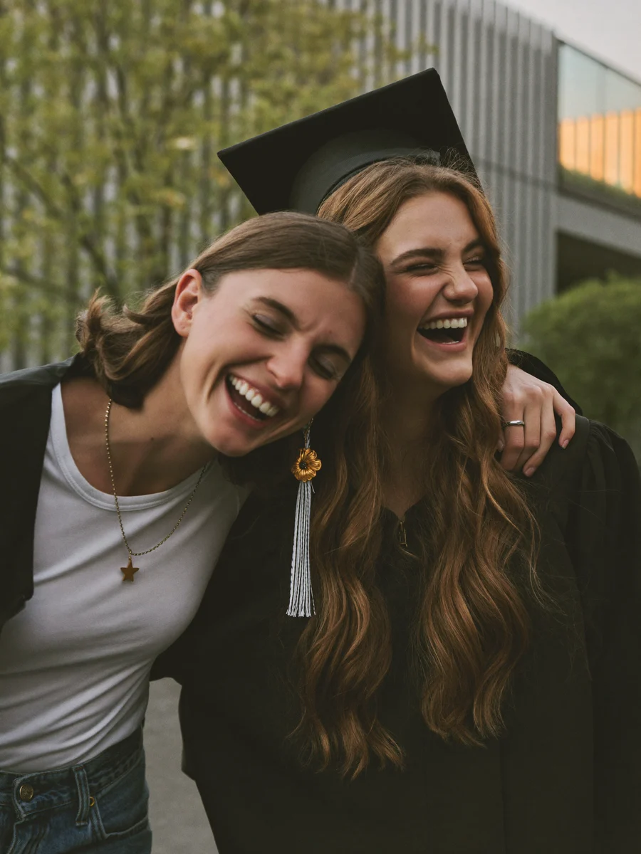 Zwei Studentinnen lachen ausgelassen zusammen im Arm, eine trägt einen Doktorhut
